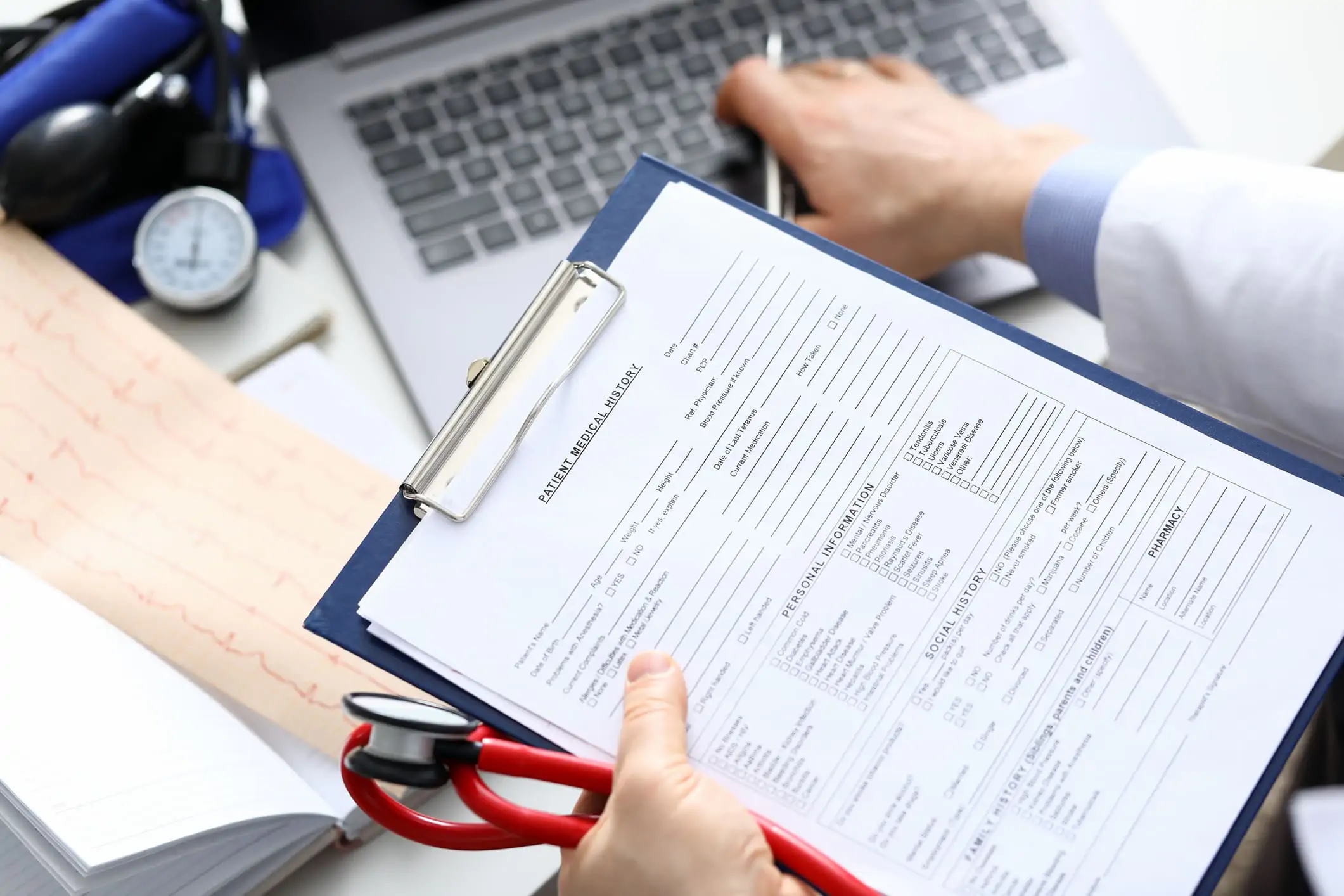 an image of a person holding a medical history form, a stethoscope, and a pen working on a laptop